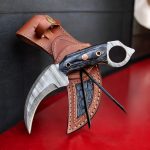 A handmade Damascus steel karambit knife with an ultra-sharp curved blade, black Pakka wood handle, and a leather sheath